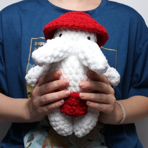 以下が含まれることがあります： 赤い帽子と赤いスカートをした、白い毛糸で編まれたぬいぐるみ。長い白い毛糸の鼻と小さな黒い目が2つある。