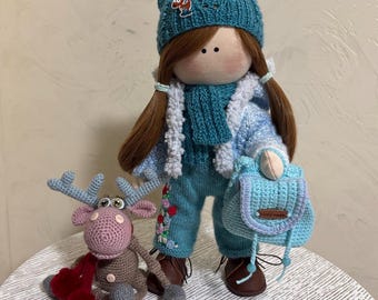 May include: A handmade doll wearing a blue knitted hat, blue sweater, and blue jeans. The doll has brown hair and is holding a blue crocheted bag. A crocheted reindeer with red hearts is sitting in front of the doll.