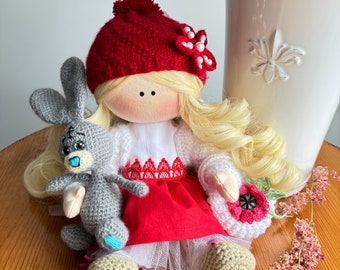 May include: A handmade doll with blonde hair wearing a red knitted hat, a white sweater, a red skirt, and a white crocheted  booties. The doll is holding a gray crocheted bunny.