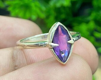 May include: A silver ring featuring a marquise-cut, deep purple gemstone. The gemstone is set in a silver band with two small, triangular accents on either side. The ring is displayed against a blurred green background.