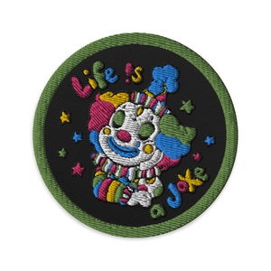 Puede incluir: Un parche bordado verde y negro con una cara de payaso y el texto "Life is a Joke". El payaso lleva un sombrero colorido y tiene una gran sonrisa.