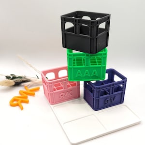 Op de afbeelding: Een stapel van vier miniatuur, kleurrijke kratten in zwart, groen, roze en blauw. Op het groene krat staat "AAA", op het roze krat "RMK" en op het blauwe krat "9V". Een witte tray staat op de voorgrond.