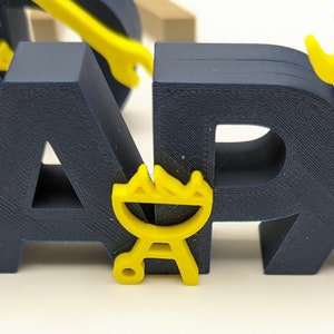 K&ouml;nnte beinhalten: Ein blaues und gelbes 3D-gedrucktes Wort "BBQ" mit einem gelben Grill-Symbol. Die Buchstaben sind dick und haben eine strukturierte Oberfl&auml;che.