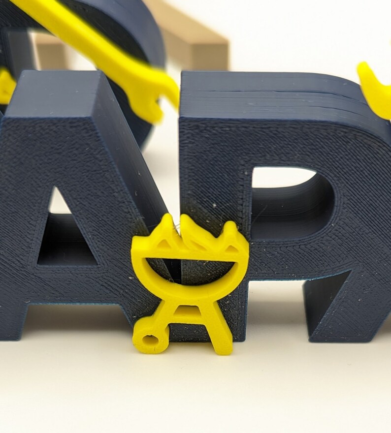 K&ouml;nnte beinhalten: Ein blaues und gelbes 3D-gedrucktes Wort "BBQ" mit einem gelben Grill-Symbol. Die Buchstaben sind dick und haben eine strukturierte Oberfl&auml;che.