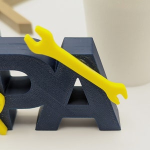 K&ouml;nnte beinhalten: Ein 3D-gedrucktes Wort "PA" in Marineblau mit einem gelben Schraubenschl&uuml;ssel oben drauf. Der Schraubenschl&uuml;ssel ist ein Symbol f&uuml;r einen Handwerker oder einen Vater.