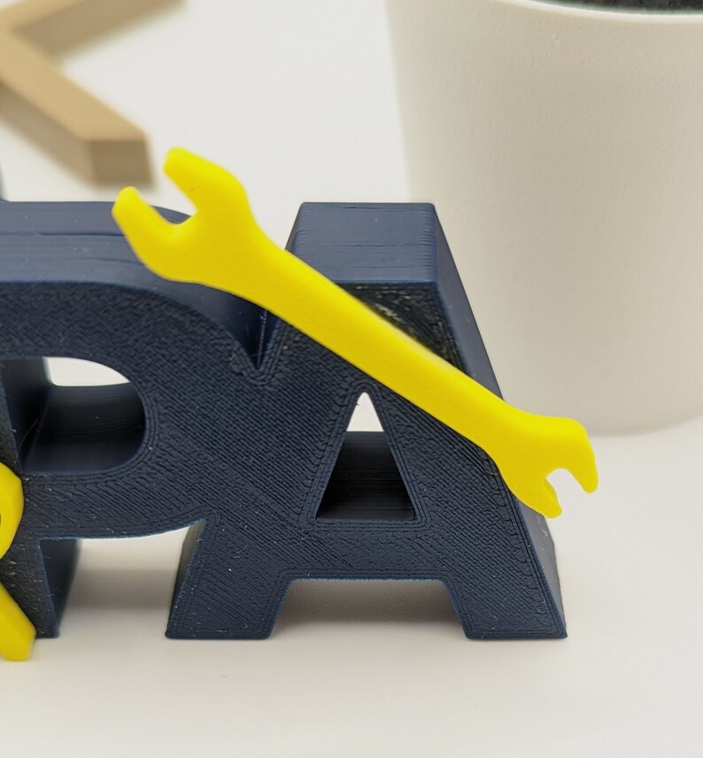K&ouml;nnte beinhalten: Ein 3D-gedrucktes Wort "PA" in Marineblau mit einem gelben Schraubenschl&uuml;ssel oben drauf. Der Schraubenschl&uuml;ssel ist ein Symbol f&uuml;r einen Handwerker oder einen Vater.