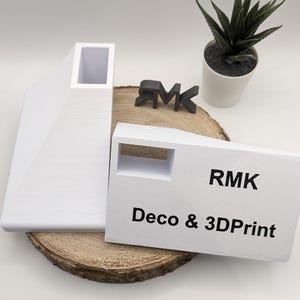 Puede incluir: Dos objetos blancos impresos en 3D, uno con una abertura rectangular y el otro con una abertura cuadrada. Los objetos est&aacute;n sobre una superficie de madera. El texto "RMK Deco & 3DPrint" est&aacute; impreso en el objeto con la abertura cuadrada.
