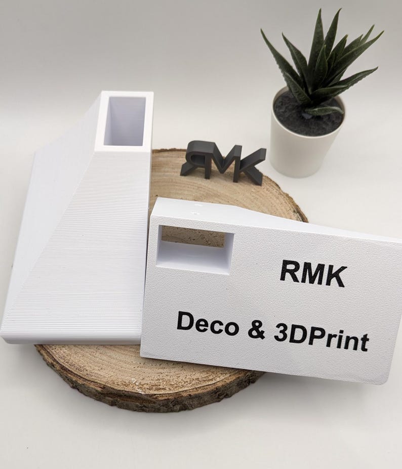 Puede incluir: Dos objetos blancos impresos en 3D, uno con una abertura rectangular y el otro con una abertura cuadrada. Los objetos est&aacute;n sobre una superficie de madera. El texto "RMK Deco & 3DPrint" est&aacute; impreso en el objeto con la abertura cuadrada.