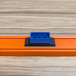Peut inclure: Une brique Lego bleue avec trois tenons est mont&eacute;e sur une base en plastique noir, qui est fix&eacute;e &agrave; une piste orange.