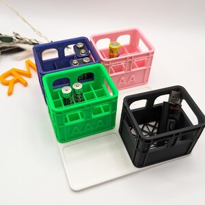 Op de afbeelding: Een verzameling kleurrijke, miniatuur kratvormige organizers. De kratten zijn blauw, roze, groen en zwart en bevatten elk verschillende items zoals batterijen en een USB-stick. De groene krat heeft de tekst "AAA", en de roze krat "AA".