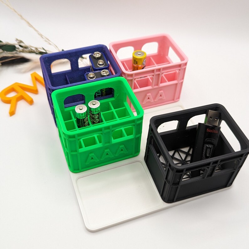 Op de afbeelding: Een verzameling kleurrijke, miniatuur kratvormige organizers. De kratten zijn blauw, roze, groen en zwart en bevatten elk verschillende items zoals batterijen en een USB-stick. De groene krat heeft de tekst "AAA", en de roze krat "AA".
