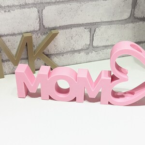 Pu&ograve; includere: Lettere in legno rosa che compongono la parola "MOM" con un cuore rosa ritagliato dietro le lettere. Le lettere sono 3D e hanno un centro vuoto.