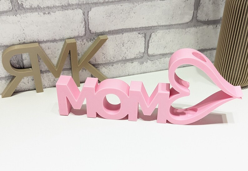 Pu&ograve; includere: Lettere in legno rosa che compongono la parola "MOM" con un cuore rosa ritagliato dietro le lettere. Le lettere sono 3D e hanno un centro vuoto.
