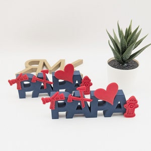 K&ouml;nnte beinhalten: Zwei blaue Holzschilder, die "PAPA" buchstabieren, mit roten Herzen und Feuerwehrhydranten-Akzenten. Die Schilder stehen auf einer wei&szlig;en Oberfl&auml;che.