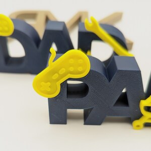 K&ouml;nnte beinhalten: Ein dreidimensionales Wort "DAD" in Marineblau mit einem gelben Videospiel-Controller auf dem "D".