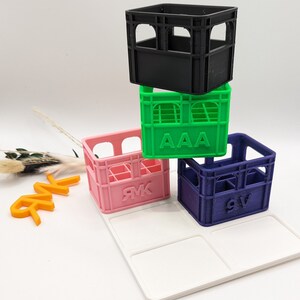 Op de afbeelding: Een stapel van vier miniatuurkratten in zwart, groen, roze en blauw, elk met een uniek ontwerp. De groene krat heeft de letters "AAA" erop, en de roze en blauwe kratten hebben letters. Ze zijn gerangschikt op een wit dienblad.