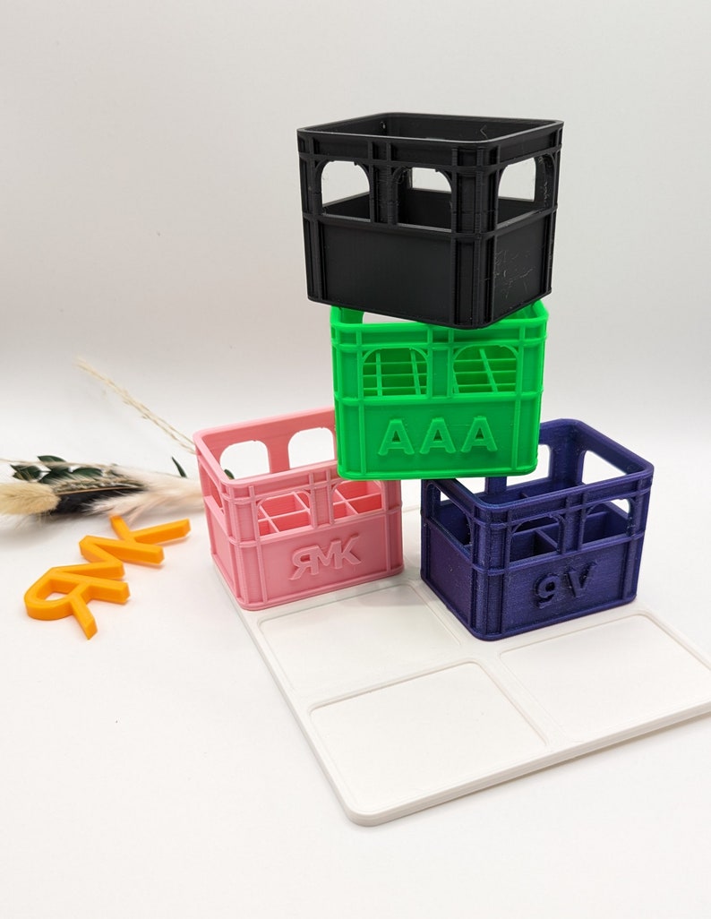 Op de afbeelding: Een stapel van vier miniatuurkratten in zwart, groen, roze en blauw, elk met een uniek ontwerp. De groene krat heeft de letters "AAA" erop, en de roze en blauwe kratten hebben letters. Ze zijn gerangschikt op een wit dienblad.