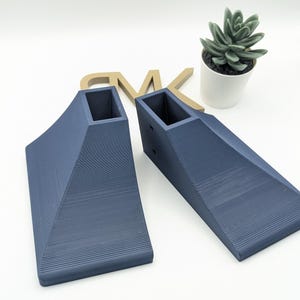 Puede incluir: Dos elevadores triangulares de forma triangular impresos en 3D de color azul oscuro con un recorte cuadrado en la parte superior. Los elevadores est&aacute;n sobre una superficie blanca.