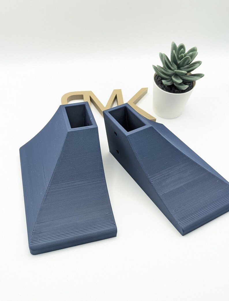 Puede incluir: Dos elevadores triangulares de forma triangular impresos en 3D de color azul oscuro con un recorte cuadrado en la parte superior. Los elevadores est&aacute;n sobre una superficie blanca.