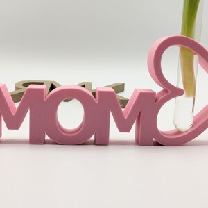 Pu&ograve; includere: Un cartello rosa MOM con un vaso a forma di cuore che contiene un unico stelo verde.