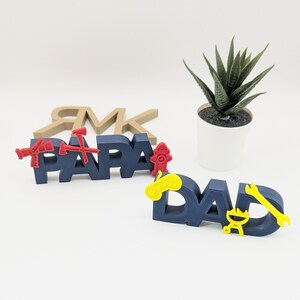 K&ouml;nnte beinhalten: Zwei blaue, 3D-gedruckte Schilder mit den Worten "Papa" und "Dad" in roten Buchstaben. Die Schilder sind mit kleinen, 3D-gedruckten Symbolen in Rot, Gelb und Schwarz verziert.