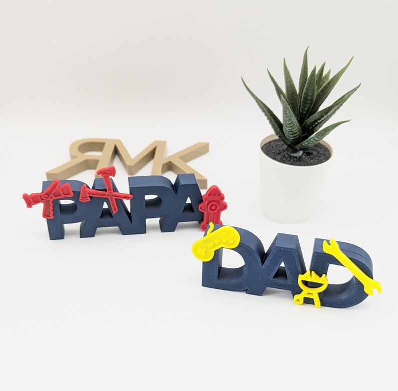 K&ouml;nnte beinhalten: Zwei blaue, 3D-gedruckte Schilder mit den Worten "Papa" und "Dad" in roten Buchstaben. Die Schilder sind mit kleinen, 3D-gedruckten Symbolen in Rot, Gelb und Schwarz verziert.