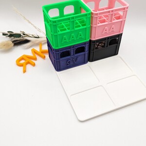 Op de afbeelding: Een stapel kleurrijke miniatuurkratten in groen, roze, marineblauw en zwart, elk met gedrukte letters. Een witte lade met vier vierkante inkepingen staat voor de kratten. Oranje letters staan links.