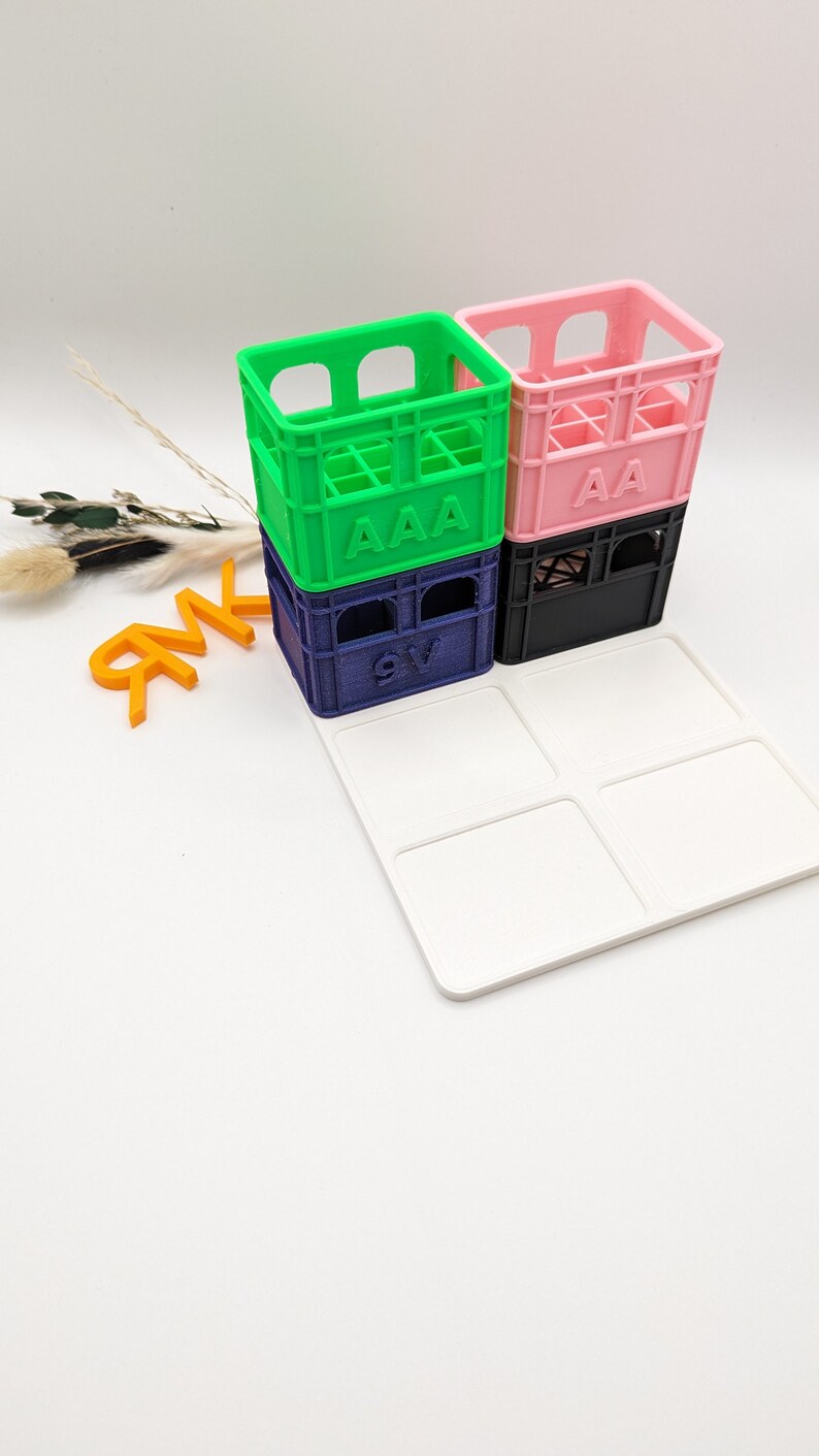 Op de afbeelding: Een stapel kleurrijke miniatuurkratten in groen, roze, marineblauw en zwart, elk met gedrukte letters. Een witte lade met vier vierkante inkepingen staat voor de kratten. Oranje letters staan links.