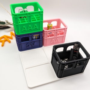 Op de afbeelding: Een verzameling kleurrijke miniatuurkratten, waaronder groene, blauwe, roze en zwarte. De groene krat is gestapeld op een blauwe krat, en de roze krat staat ernaast. De kratten zijn gemarkeerd met "AAA", "9V" en "AA". Een zwarte krat bevat een USB-stick.