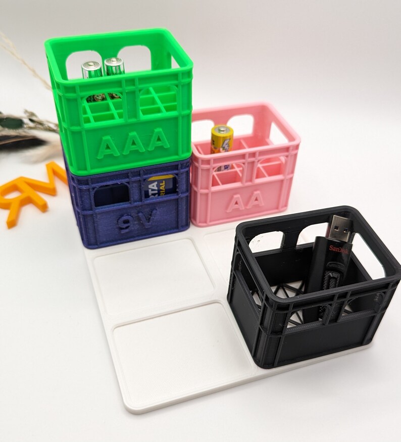 Op de afbeelding: Een verzameling kleurrijke miniatuurkratten, waaronder groene, blauwe, roze en zwarte. De groene krat is gestapeld op een blauwe krat, en de roze krat staat ernaast. De kratten zijn gemarkeerd met "AAA", "9V" en "AA". Een zwarte krat bevat een USB-stick.