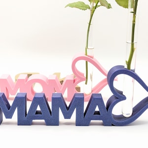 Pu&ograve; includere: Un cartello in legno blu che scrive "MAMA" con due vasi di fiori a forma di cuore rosa e blu con provette di vetro trasparente per contenere i fiori.