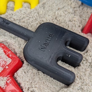 Pode incluir: Ancinho de pl&aacute;stico preto com a palavra "Name" impressa no cabo. O ancinho est&aacute; em um monte de areia.