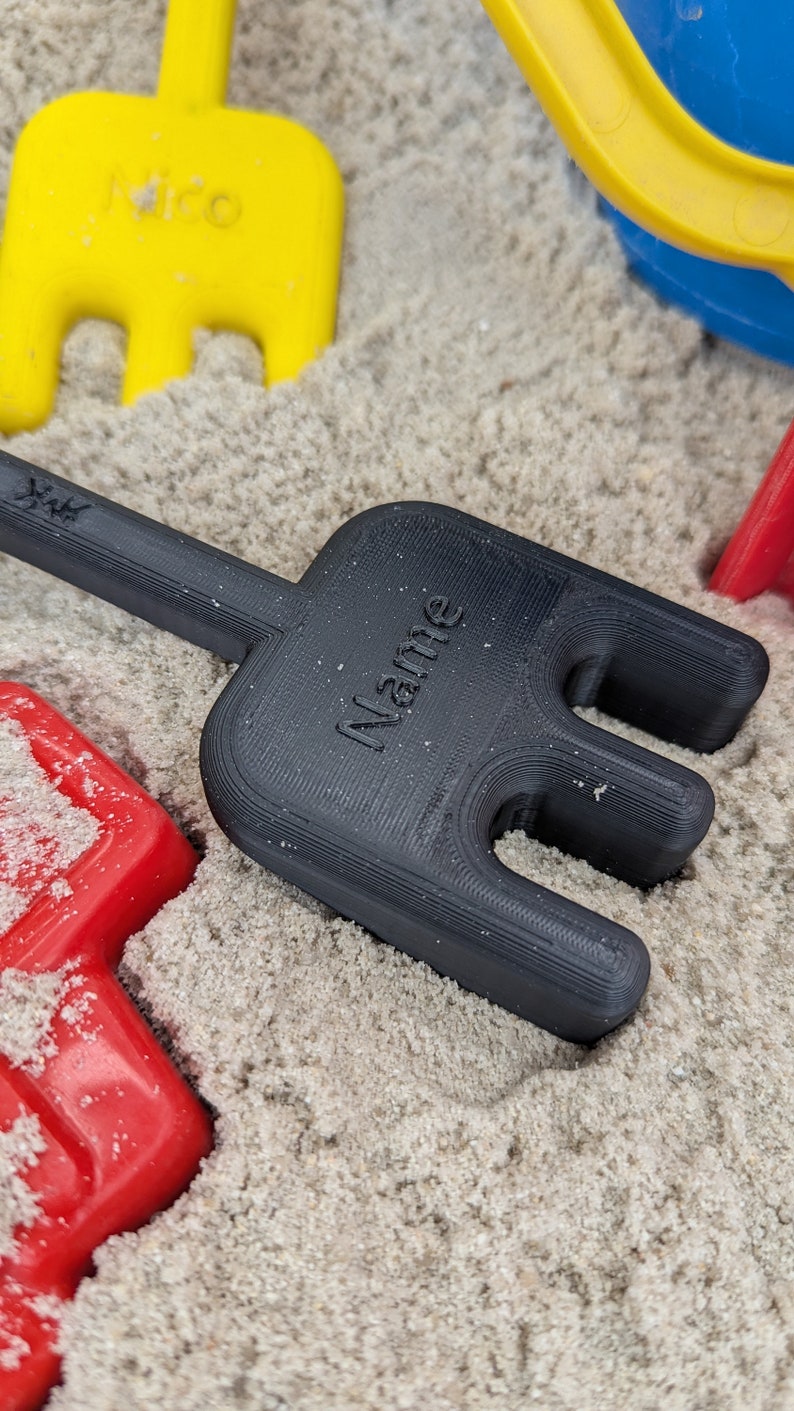 Pode incluir: Ancinho de pl&aacute;stico preto com a palavra "Name" impressa no cabo. O ancinho est&aacute; em um monte de areia.