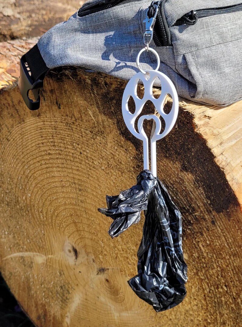 以下が含まれることがあります： 白いプラスチック製の犬の糞袋ディスペンサーで、足跡のデザインが施されています。ディスペンサーは、銀色のクリップが付いた黒い袋に取り付けられています。