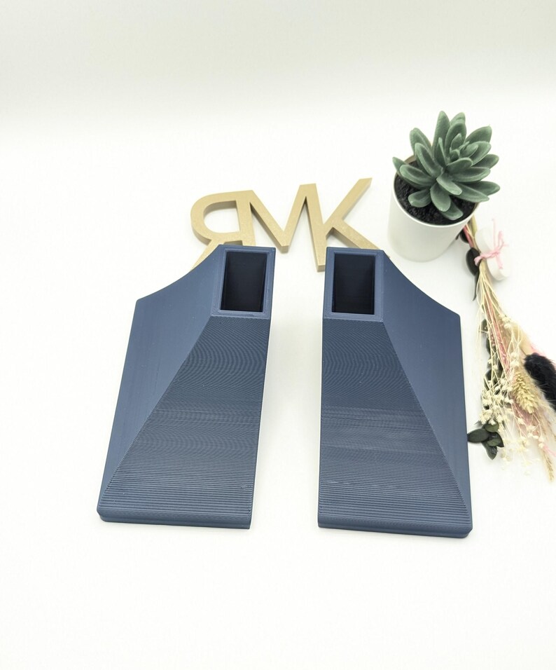 Puede incluir: Dos soportes de estante triangulares de color azul oscuro, impresos en 3D, con un recorte cuadrado en el centro.