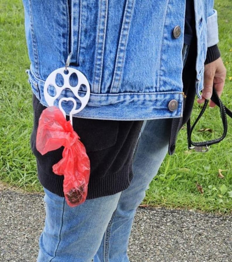 以下が含まれることがあります： 人のベルトループに付けられた、足跡のデザインが施された白いプラスチック製の犬の糞袋ディスペンサー。赤いプラスチック製の袋がディスペンサーからぶら下がっています。