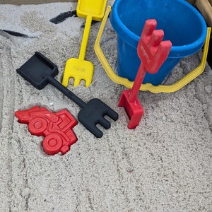 Pode incluir: Um conjunto de brinquedos de pl&aacute;stico coloridos para crian&ccedil;as para brincar em uma caixa de areia. O conjunto inclui um caminh&atilde;o de brinquedo vermelho, uma p&aacute; amarela, um balde azul e dois ancinhos pretos. Os brinquedos est&atilde;o em uma cama de areia branca.