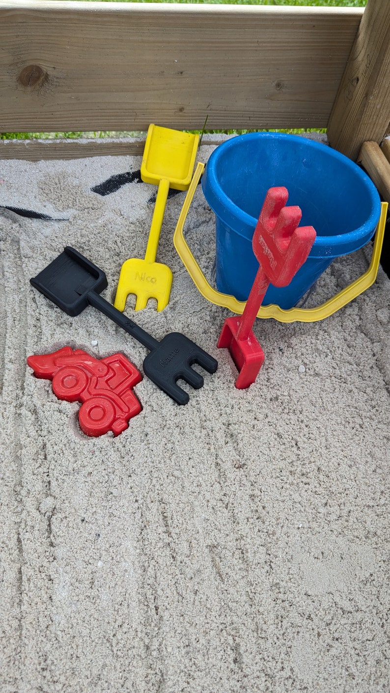 Pode incluir: Um conjunto de brinquedos de pl&aacute;stico coloridos para crian&ccedil;as para brincar em uma caixa de areia. O conjunto inclui um caminh&atilde;o de brinquedo vermelho, uma p&aacute; amarela, um balde azul e dois ancinhos pretos. Os brinquedos est&atilde;o em uma cama de areia branca.