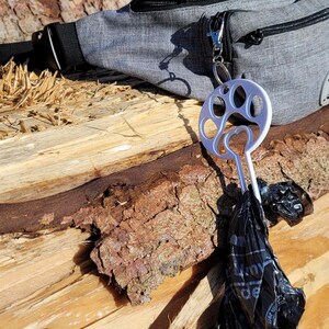 以下が含まれることがあります： 灰色のウエストバッグに、銀色の足跡の形をしたディスペンサーが取り付けられています。黒いビニール袋がディスペンサーからぶら下がっています。