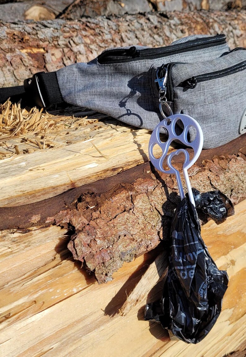 以下が含まれることがあります： 灰色のウエストバッグに、銀色の足跡の形をしたディスペンサーが取り付けられています。黒いビニール袋がディスペンサーからぶら下がっています。