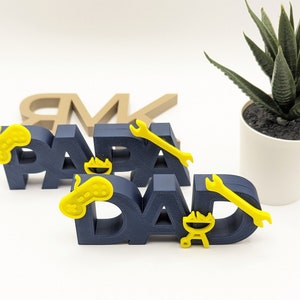 K&ouml;nnte beinhalten: Zwei blaue, 3D-gedruckte Schilder, auf denen "Papa" und "Papa" steht. Die Schilder haben gelbe Akzente, darunter einen Videospiel-Controller, einen Schraubenschl&uuml;ssel und einen Grill.