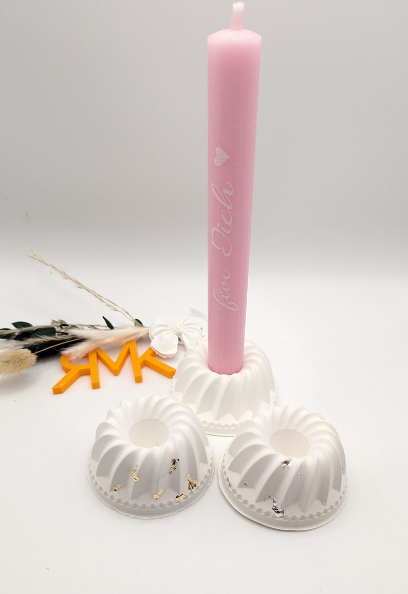 Peut inclure: Trois bougeoirs blancs avec une bougie rose dans le bougeoir central. La bougie porte l'inscription "f&uuml;r dich". Les bougeoirs ont un motif en spirale et des accents dor&eacute;s et argent&eacute;s.