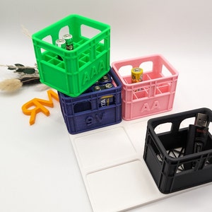 Op de afbeelding: Een verzameling kleine, kleurrijke kratten, elk met verschillende soorten batterijen en een USB-stick. De kratten zijn groen, roze, blauw en zwart, met labels "AAA", "AA" en "9V". De items zijn gerangschikt op een witte tray.