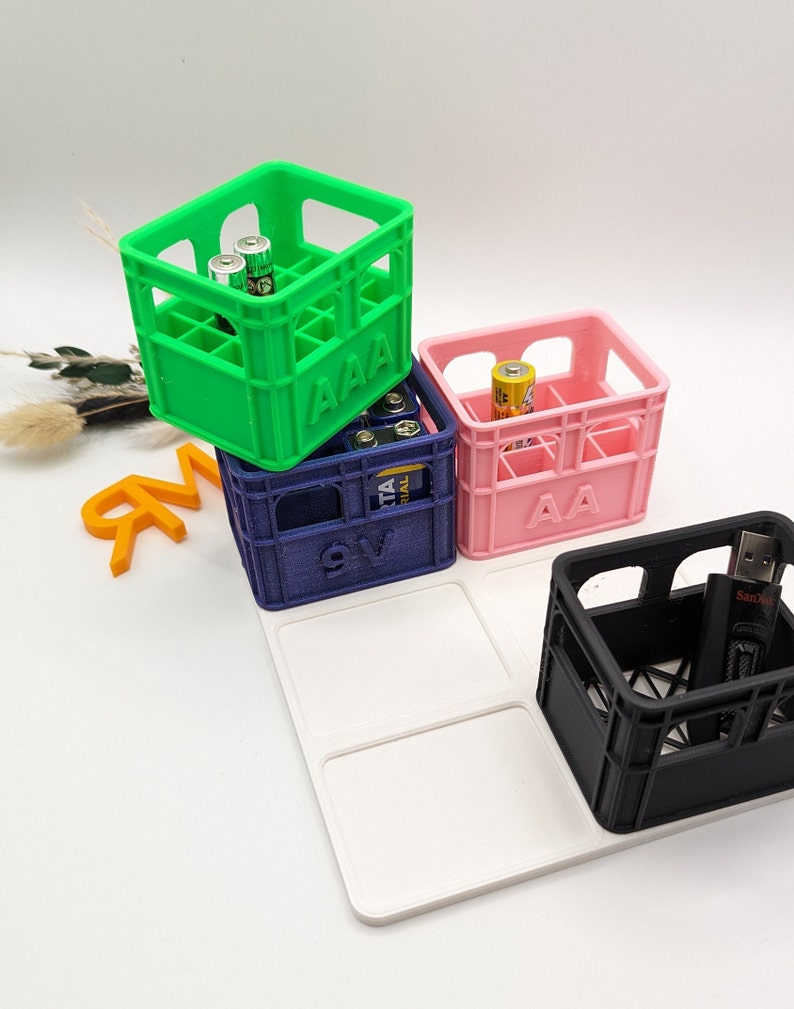 Op de afbeelding: Een verzameling kleine, kleurrijke kratten, elk met verschillende soorten batterijen en een USB-stick. De kratten zijn groen, roze, blauw en zwart, met labels "AAA", "AA" en "9V". De items zijn gerangschikt op een witte tray.