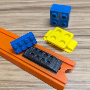 Peut inclure: Gros plan sur une piste de voiture jouet avec une pi&egrave;ce en plastique noire, un bloc Lego bleu et un bloc Lego jaune. La piste est orange et a un bord sur&eacute;lev&eacute;.
