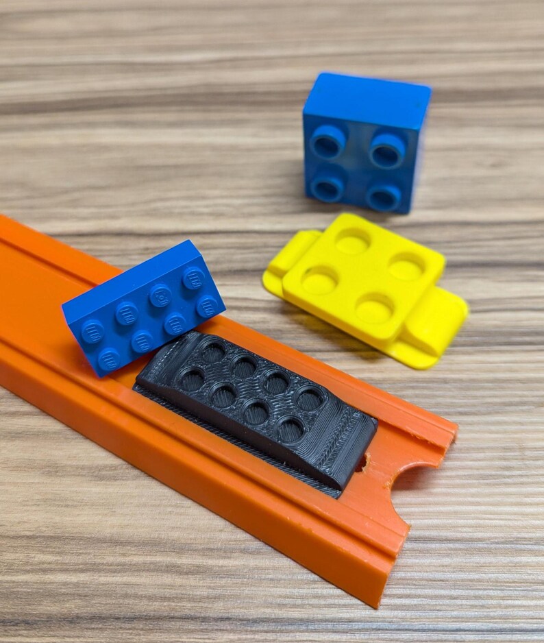 Peut inclure: Gros plan sur une piste de voiture jouet avec une pi&egrave;ce en plastique noire, un bloc Lego bleu et un bloc Lego jaune. La piste est orange et a un bord sur&eacute;lev&eacute;.