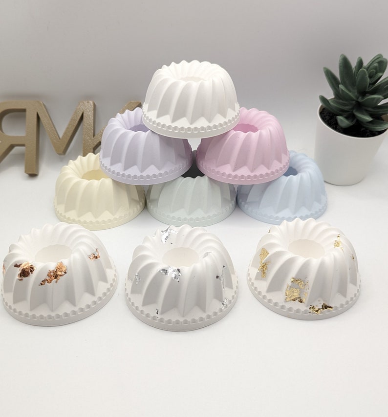 Peut inclure: Un ensemble de huit petits bougeoirs en forme de g&acirc;teau, de couleur blanche. Certains sont blancs unis, tandis que d'autres ont des accents de feuilles d'or ou d'argent. Les bougeoirs sont dispos&eacute;s en forme de pyramide.