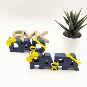 K&ouml;nnte beinhalten: Zwei blaue, 3D-gedruckte W&ouml;rter, "DAD" und "PAPA", mit gelben Akzenten. Die Akzente umfassen einen Schraubenschl&uuml;ssel, einen Videospiel-Controller und einen Grill.