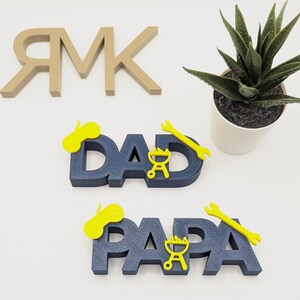 K&ouml;nnte beinhalten: Zwei blaue Holzschilder mit den Worten "DAD" und "PAPA" in wei&szlig;en Buchstaben. Jedes Schild hat einen gelben Schraubenschl&uuml;ssel, einen Grill und einen gelben Kreis oben drauf.