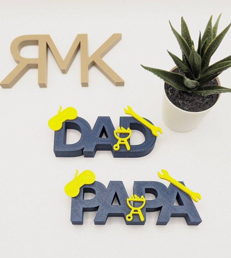 K&ouml;nnte beinhalten: Zwei blaue Holzschilder mit den Worten "DAD" und "PAPA" in wei&szlig;en Buchstaben. Jedes Schild hat einen gelben Schraubenschl&uuml;ssel, einen Grill und einen gelben Kreis oben drauf.